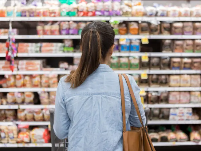 A woman stands with a shopping cart in front of a shelf full of food in a grocery store.